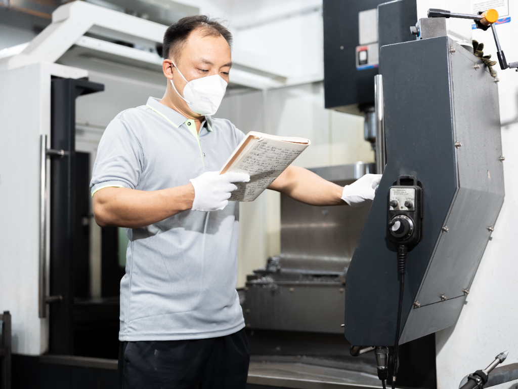 A technician in a mask and gloves inspects notes while operating a CNC machine for mould making.