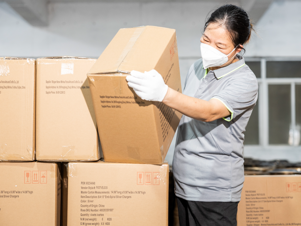 A factory worker in a mask and gloves lifts a box of products for shipping.