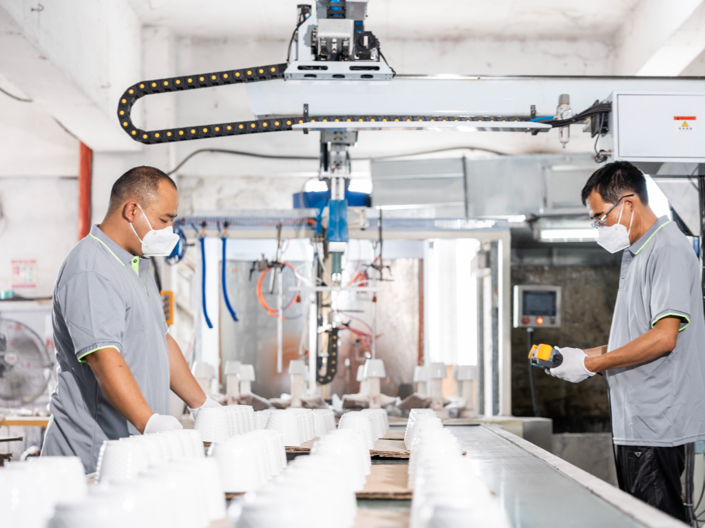 Two factory workers in masks and gloves supervise plastic flower pots on a spray painting conveyor belt.