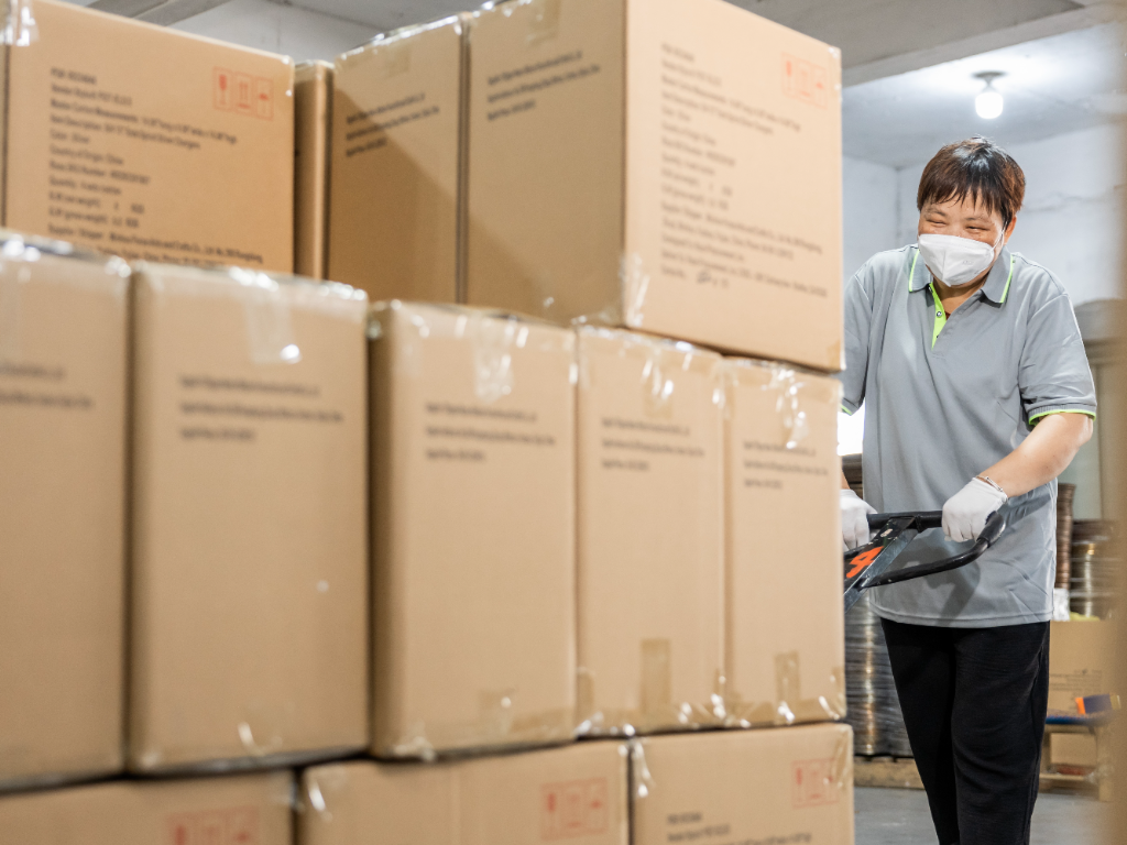 A factory worker uses a pallet jack to move large stacks of packaged boxes.