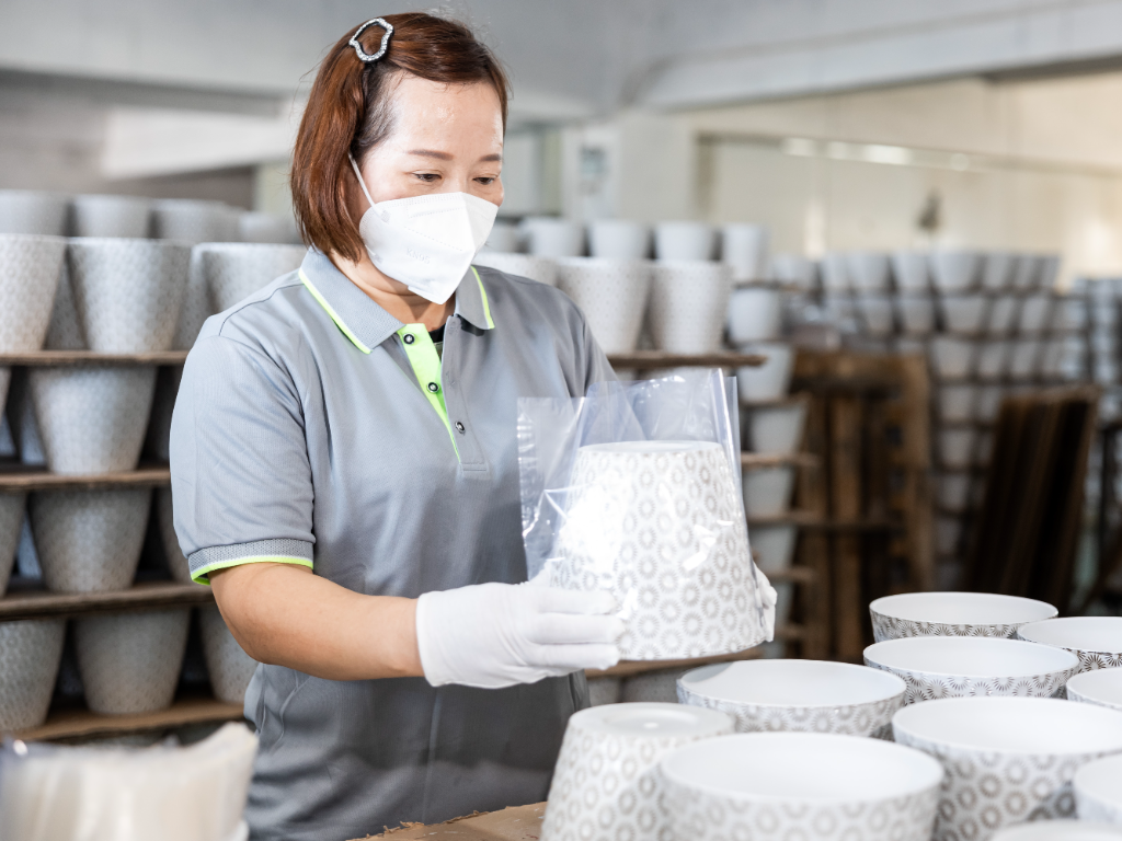 A factory worker in a mask and gloves carefully places a patterned plastic flower pot into a protective bag.